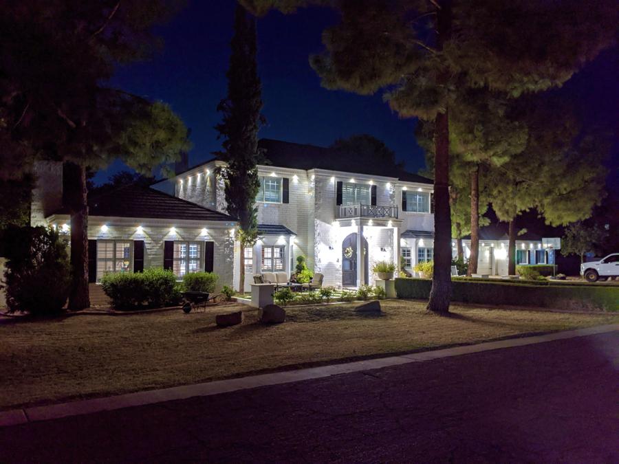 A two-story white brick home at night with warm white permanent lights along every roofline